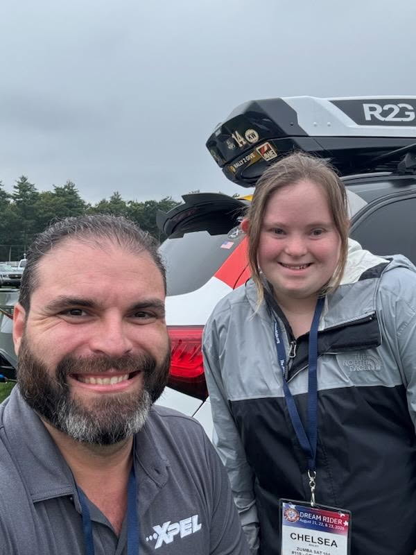 A smiling man with a beard and a woman with Down syndrome pose for a selfie in front of a car with a roof rack, wearing casual attire and event badges.