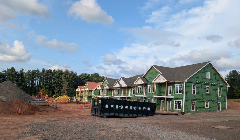 Construction site featuring green multi-family buildings in progress, with a large dumpster and piles of gravel in the foreground, under a partly cloudy sky.