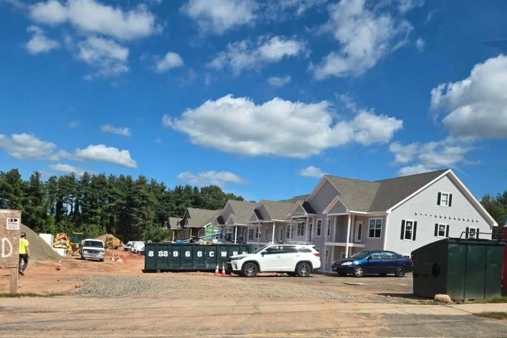 A construction site featuring new residential buildings, parked vehicles, and construction equipment under a clear blue sky with fluffy clouds.
