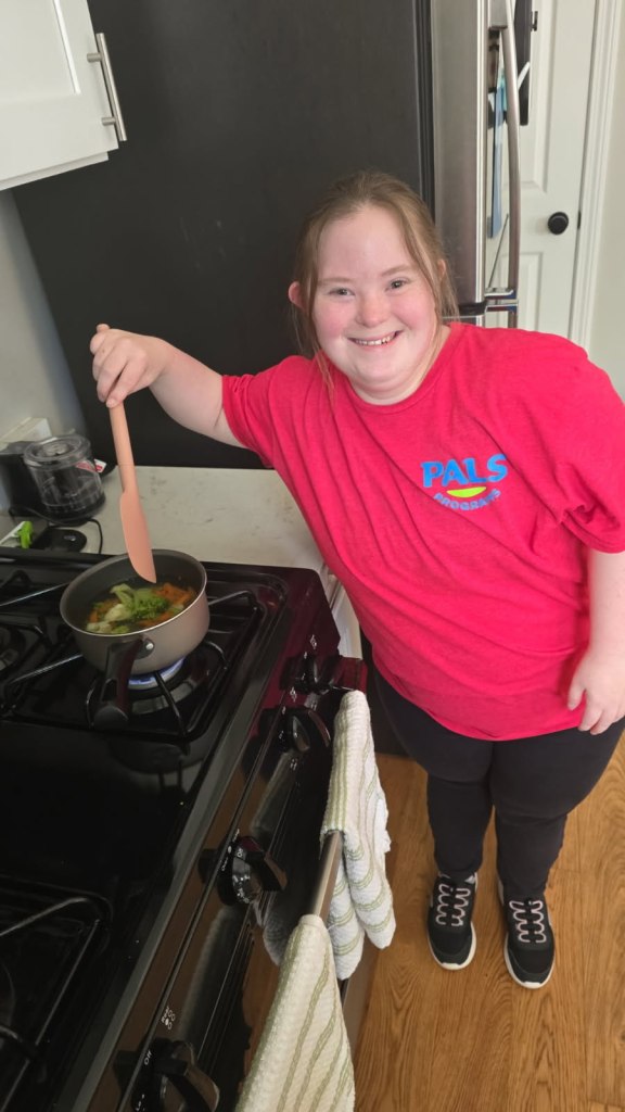 A young person in a red shirt stands at a stove, happily stirring vegetables in a small pot with a wooden spoon.