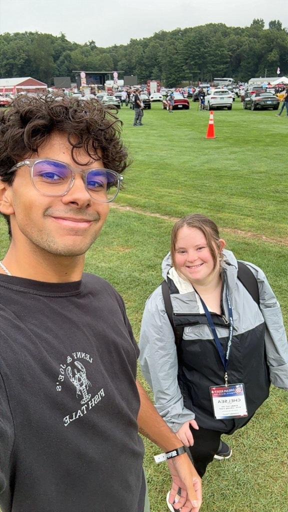 Two young people smiling for a selfie at an outdoor event, with a grassy field and cars in the background.