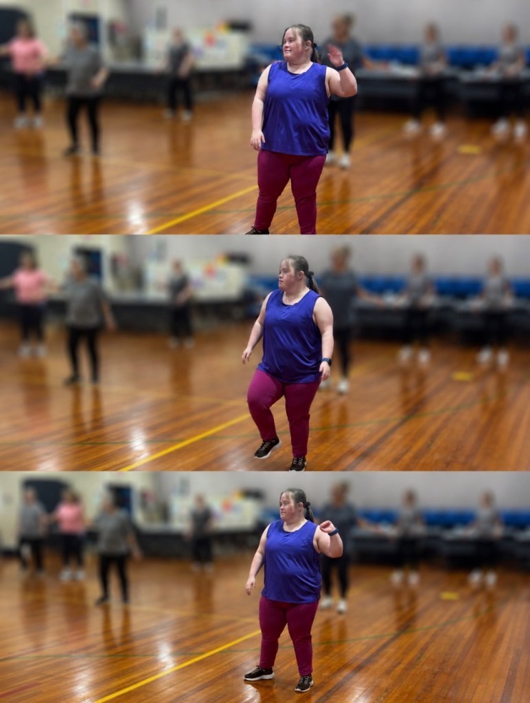 A person with Down syndrome is participating in a dance class, wearing a blue top and maroon pants, standing on a wooden floor surrounded by other dancers in a gym setting.