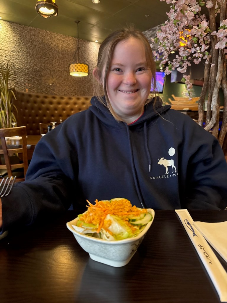A girl sitting at a restaurant table, smiling while holding a fork. In front of her, there is a bowl of salad topped with shredded carrots and cucumber slices.