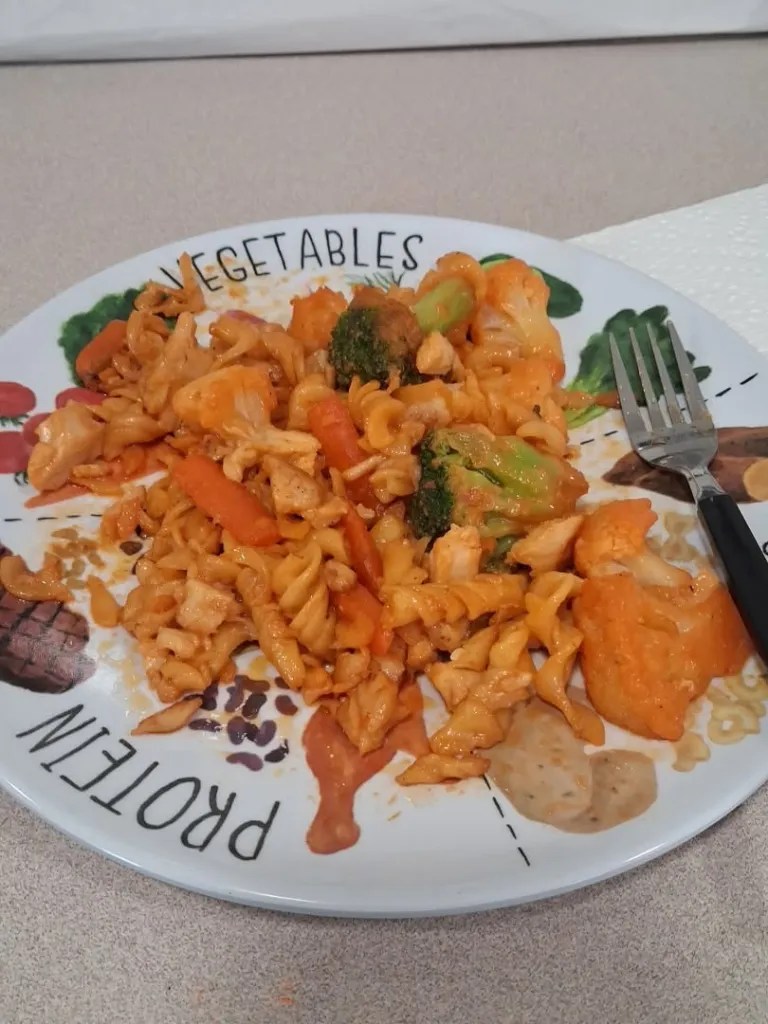 A plate of pasta with vegetables including broccoli and carrots, topped with a creamy sauce. A fork is placed beside the plate.