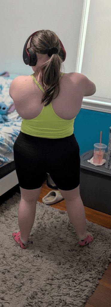 A person wearing a bright yellow tank top and black shorts, viewed from behind, is standing in a room with a rug and other furniture. They are wearing headphones and appear to be engaged in a workout or stretching activity.