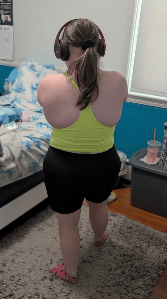A person wearing a bright yellow tank top and black shorts, standing in a room with headphones on, facing away from the camera. The background includes a bed with blue bedding, a rug, and a container on the floor.